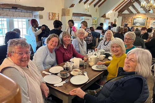 A large group of NHS Retirement Fellowship members smiling at the camera while seated around a table in a busy café, with other tables also filled with fellow NHS Retirement Fellowship members enjoying the gathering.