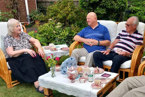 Four NHS Retirement Fellowship members—one woman and three men—seated in a garden around a low table, enjoying afternoon tea and chatting together.
