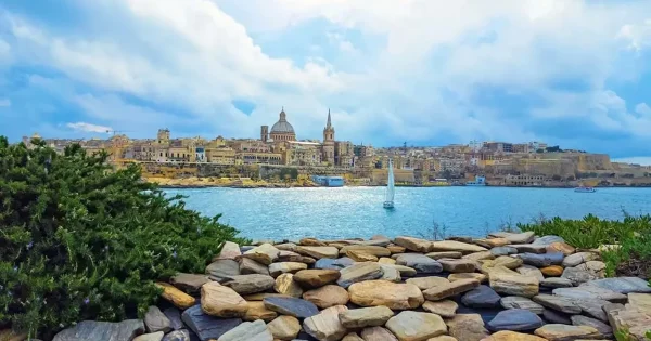 View of Valletta, Malta, from a distant rocky shore, showcasing the city’s historic buildings and harbor under a clear sky.